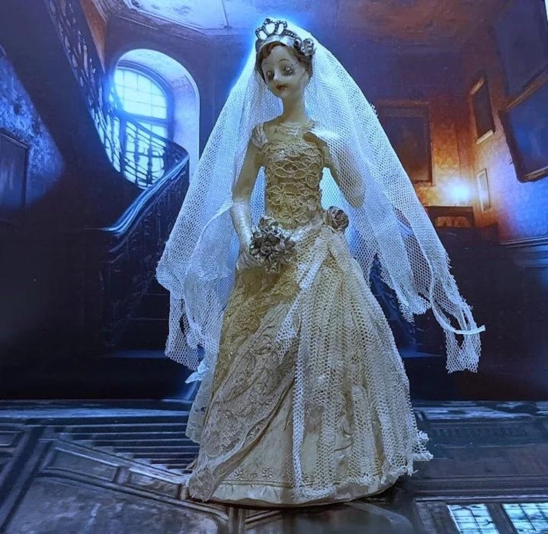 May include: A porcelain doll bride figurine wearing a white lace wedding dress and veil. The doll is standing on a wooden floor with a staircase in the background.