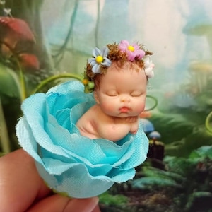 Può includere: Statuina di un bambino addormentato in miniatura, adagiato in una rosa azzurra. Il bambino indossa una corona floreale con fiori rosa, gialli e bianchi. La statuina è un pezzo dettagliato e fatto a mano, perfetto per giardini fatati o case di bambole.