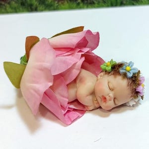Puede incluir: Figura de un bebé durmiente acurrucado dentro de una flor rosa. El bebé tiene una corona floral con detalles azules y verdes. Los pétalos de la flor son de color rosa claro y verde.