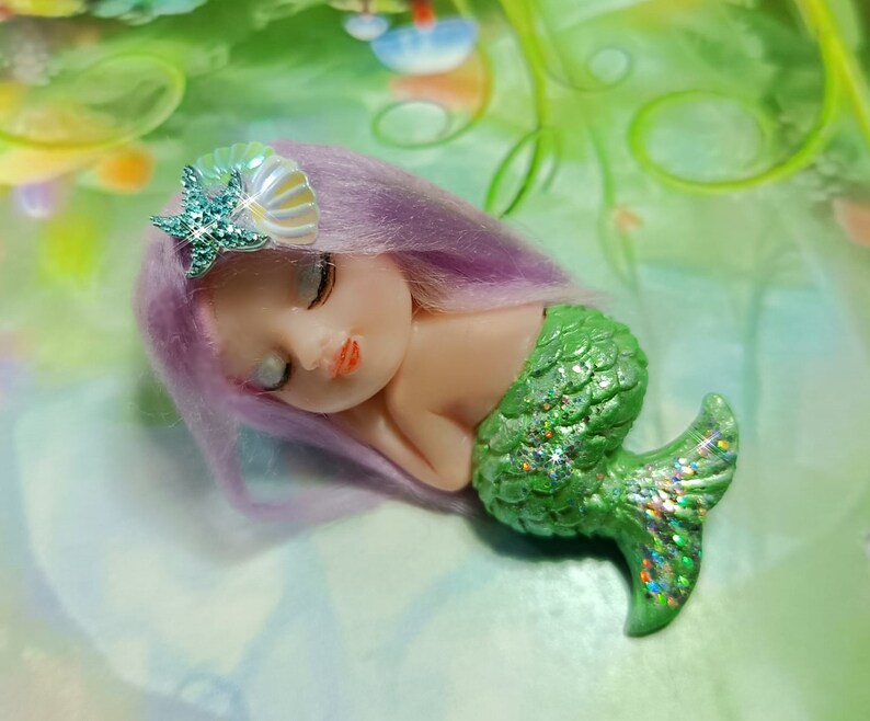 May include: A small, sleeping mermaid doll with purple hair and a green, glittery tail. She is wearing a seashell and starfish hair clip.