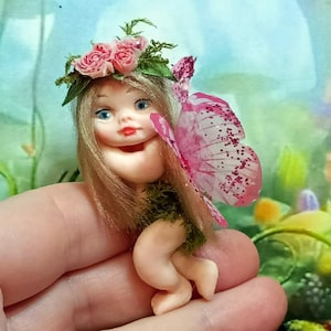 Könnte beinhalten: Eine kleine Porzellan-Feenpuppe mit rosa Flügeln und einer Blumenkrone. Die Fee hält sich das Kinn und hat einen süßen Gesichtsausdruck.