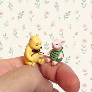 May include: Two miniature figurines of a yellow bear and a pink piglet. The yellow bear has a brown string tied around its neck. The pink piglet wears a green shirt with dark green horizontal stripes. Both figures are in a seated position.