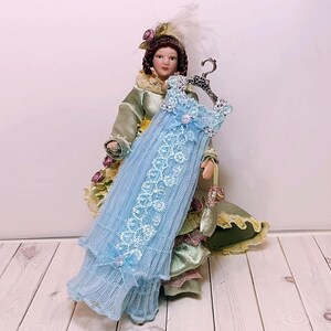 May include: A miniature doll dressed in a green and gold gown, holding a light blue dress on a silver hanger. The dress is adorned with white lace and floral accents. The doll has brown hair and is wearing a feathered hat.