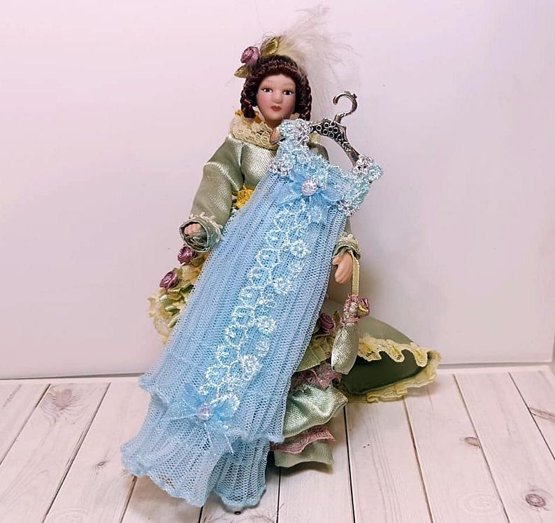 May include: A miniature doll dressed in a green and gold gown, holding a light blue dress on a silver hanger. The dress is adorned with white lace and floral accents. The doll has brown hair and is wearing a feathered hat.