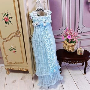 May include: A light blue doll dress with white lace detailing, hanging on a silver hanger. The dress features a delicate, layered design with a ruffled hem and a bow. The dress is displayed near a vintage-style cabinet.