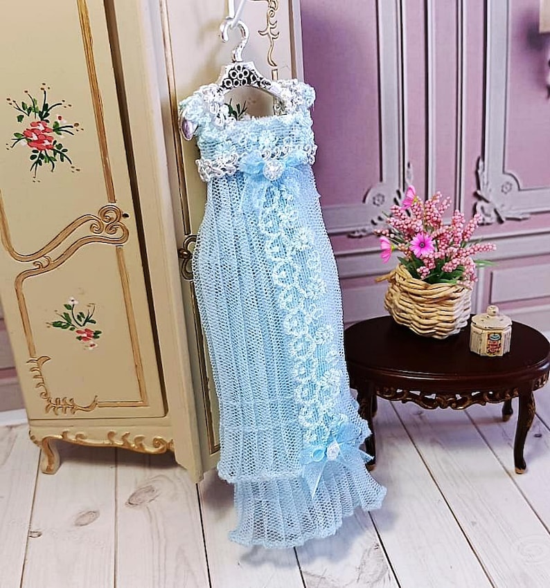 May include: A light blue doll dress with white lace detailing, hanging on a silver hanger. The dress features a delicate, layered design with a ruffled hem and a bow. The dress is displayed near a vintage-style cabinet.