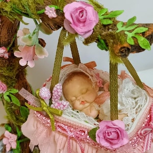 May include: A sleeping baby doll in a pink and white swing made of fabric and lace. The swing is suspended from a branch with green moss and pink flowers.