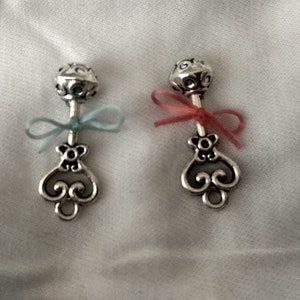 Puede incluir: Dos abalorios de sonajero de bebé de plata con detalles decorativos. Un abalorio tiene una cinta azul claro y el otro tiene una cinta rosa.