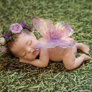 May include: A sleeping baby fairy figurine with a floral crown and translucent purple wings. The baby is lying on its stomach with its eyes closed. The wings are a gradient of pink and purple.