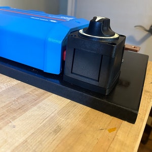 May include: A blue electrical component with a black rotary switch and a copper terminal. The component is mounted on a black base, resting on a wooden surface. The switch has a white arrow indicator.