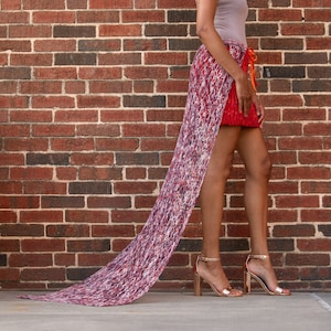 May include: A woman wearing a red skirt with a long, flowing, multi-colored scarf attached to the back. She is standing in front of a brick wall and smiling.