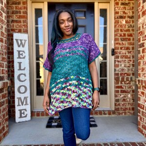 May include: A vibrant, short-sleeved crochet top in shades of purple, teal, and green. The top has a loose fit and an open-weave design. The person is wearing blue jeans. A wooden sign that says "WELCOME" is visible.