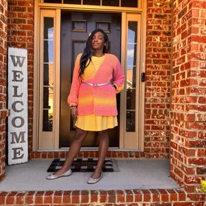 May include: A woman wearing a yellow dress and a pink and yellow cardigan. The cardigan is belted at the waist with a silver belt. The background includes a brick wall, a door, and a welcome sign.