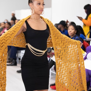 May include: A woman wearing a black dress with a gold chain belt and a crocheted mustard yellow shawl. She is walking down a runway.