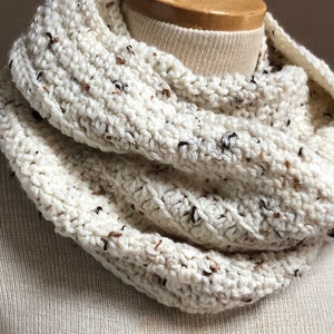 May include: A white crocheted infinity scarf with brown flecks. The scarf is folded and displayed on a white mannequin.