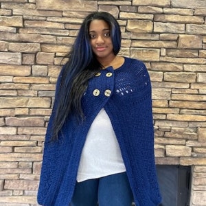 May include: A dark blue crocheted cape with three large wooden buttons. The cape has a textured pattern and is worn over a white top and blue jeans. The background is a brick wall.