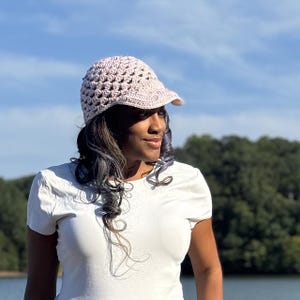 May include: A light pink crocheted cap with a visor. The cap has an open weave design, allowing for ventilation. The cap is worn by a person with dark hair and a white shirt, set against a backdrop of trees and a blue sky.