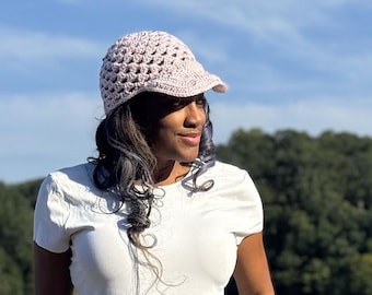 Può includere: Un berretto all'uncinetto rosa chiaro con visiera. Il berretto presenta un motivo a trama aperta. Il berretto è indossato da una persona con capelli scuri e una camicia bianca, sullo sfondo di alberi e cielo blu.