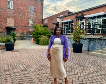 May include: A person wearing a purple crochet shrug over a white top and a long, beige skirt. The person is standing in front of a brick building with a sign that reads "Naked Spirits Distillery".