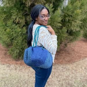 May include: A blue and teal crocheted handbag with matching straps. The bag has a rounded shape and a textured appearance. The bag is being carried over the shoulder. The background includes green foliage and a grassy area.