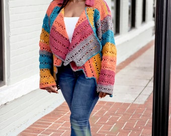 May include: A woman wearing a multi-colored crochet cardigan with a white tank top and blue jeans. She is walking on a brick sidewalk.