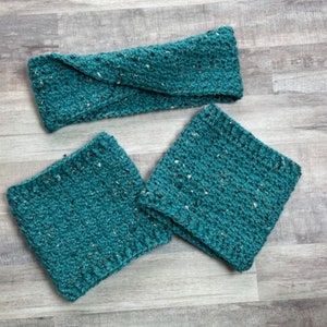 May include: A teal-colored crochet headband and two matching wrist cuffs. The items are made of a textured yarn with flecks of white and tan. The headband is twisted at the front. The items are displayed on a light gray wooden surface.