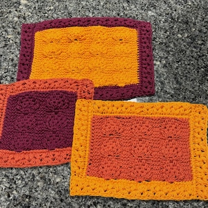 Puede incluir: Tres paños de cocina de ganchillo en naranja, rojo y morado con fondo blanco. Los paños de cocina están hechos con un patrón de punto simple y tienen un borde alrededor de los bordes.