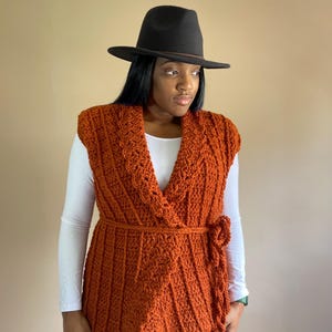 May include: A rust-colored crochet vest with a wrap style and a tie belt. The vest has a scalloped hem and is worn over a white long-sleeved shirt. The person is wearing a black fedora hat.