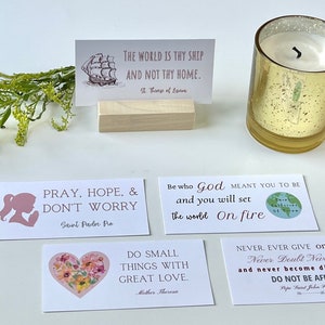 May include: An arrangement of inspirational cards with quotes, a gold candle holder, and greenery. The cards feature quotes like "Pray, Hope, & Don't Worry" and "Do Small Things With Great Love."