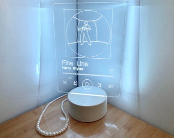 May include: White acrylic night light with a silhouette of a person dancing inside a circle. The light has a music player design with the text "Fine Line Harry Styles" and play, pause, and skip buttons.