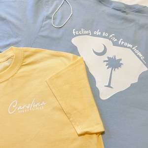 May include: Two t-shirts, one light blue and one yellow, with white text. The light blue t-shirt has a white outline of the state of South Carolina with a palm tree and crescent moon. The text on the light blue t-shirt reads "feeling oh so far from home...". The yellow t-shirt has the text "Carolina Handcrafted" in white.