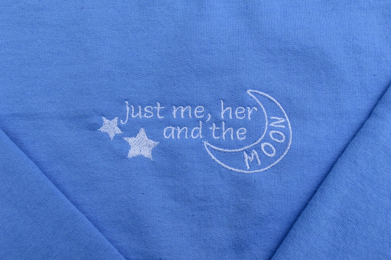 Puede incluir: Una sudadera azul claro con bordado blanco que dice "Just me, her and the moon" con dos estrellas y una luna creciente.