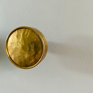 May include: A round, hammered brass cabinet knob with a gold finish. The knob has a circular design with a textured surface and a smooth, rounded edge. The hardware is set against a white background.