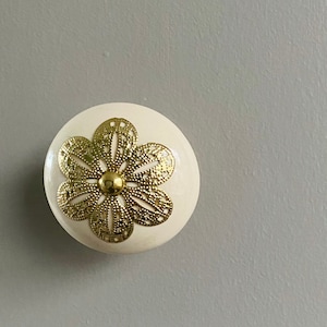 May include: A white ceramic knob with a gold metal flower design. The knob is round and has a smooth surface.