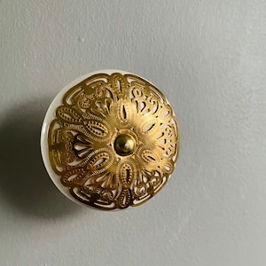 May include: White ceramic knob with a gold filigree design. The knob is round and has a raised centre.