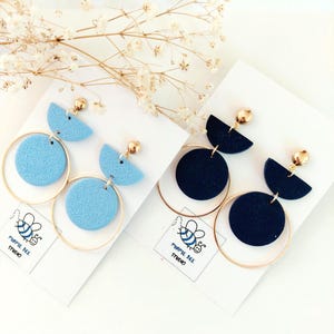 May include: Two pairs of dangle earrings on white display cards labelled Purple Bee Studio. One pair is light blue, the other is dark navy blue. Each earring features a textured semi-circle, a textured circle, and a thin gold-tone metal hoop.