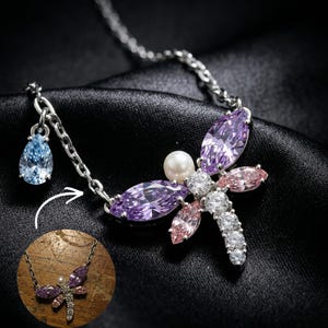 May include: A silver necklace featuring a dragonfly design, adorned with purple, pink, and clear crystals. A pearl is set in the centre. A blue, teardrop-shaped crystal pendant hangs from the chain.