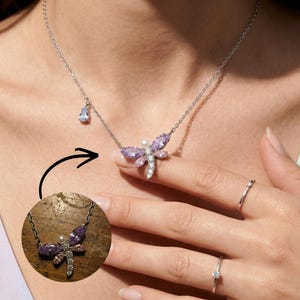 May include: A silver dragonfly necklace with purple and pink gemstone wings and a pearl body. The necklace has a small blue teardrop charm. Two silver rings are also visible.
