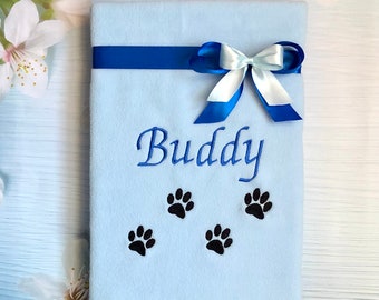 Pode incluir: Uma manta de lã azul claro com uma fita azul amarrada em um laço. A manta é bordada com o nome "Buddy" e quatro pegadas pretas.