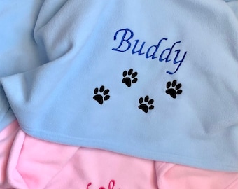 Pode incluir: Duas mantas de lã personalizadas para animais de estimação. Uma é azul claro com o nome "Buddy" e pegadas bordadas em preto. A outra é rosa com o nome "Chloe" e pegadas bordadas em preto.