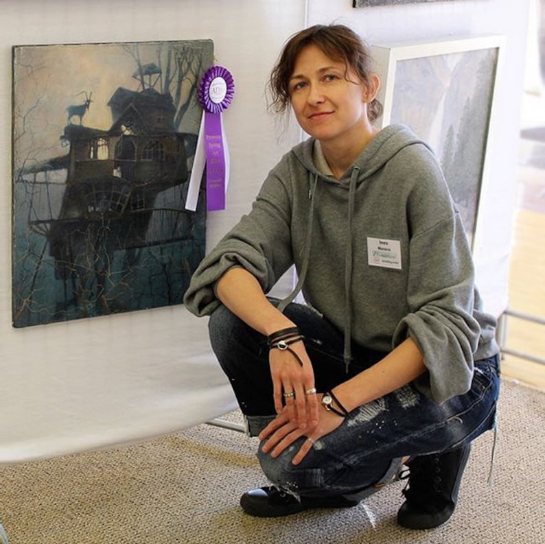 Puede incluir: Una mujer con una sudadera gris y pantalones vaqueros azules est&aacute; arrodillada frente a una pintura. La pintura representa una casa en un &aacute;rbol con un ciervo en una rama. Una cinta morada con un sello dorado est&aacute; unida a la pintura.