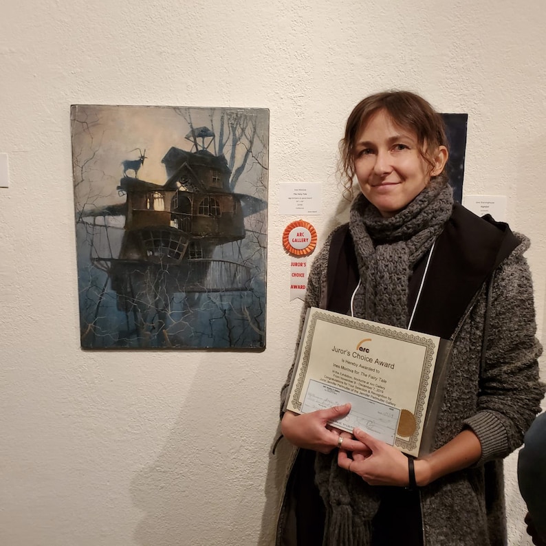 Puede incluir: Una mujer sosteniendo un certificado que dice "Junior's Choice Award" otorgado a "The Mountain for The Fairy Tale" por "Art Gallery" en "2018". El certificado est&aacute; enmarcado con un borde dorado. La mujer lleva una bufanda gris y una chaqueta negra. Est&aacute; de pie frente a una pintura de una casa en un &aacute;rbol en el bosque.