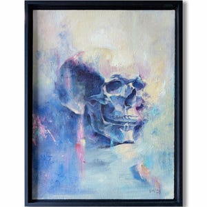 May include: An oil painting of a skull in a black frame. The skull is rendered in shades of blue and purple, with a soft, ethereal quality. The background features a blend of light blue, pink, and white hues, creating a dreamlike atmosphere.