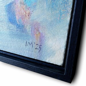 May include: An abstract oil painting with a palette of blues, purples, and creams, framed in a black wooden frame. The artwork features visible brushstrokes and the artist's initials and year, "IM '23", in the lower right corner. The painting is angled, showcasing the textured canvas.