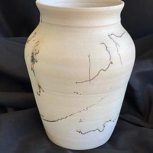 May include: A cream-colored ceramic vase with a wide body and a slightly flared rim. The vase features dark, organic-shaped markings that add a unique, artistic touch. The vase is suitable for home decor.