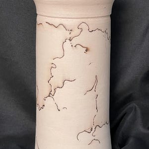 May include: A cylindrical, off-white ceramic vase with a slightly flared top. The vase features dark brown, organic-shaped markings across its surface. The design gives the vase a unique, handcrafted appearance. The vase is suitable for home decor.