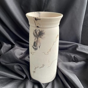 May include: A white ceramic vase with a black abstract design. The vase has a cylindrical shape and a slightly flared rim.