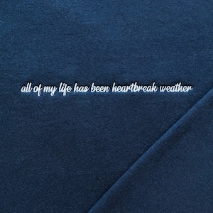 May include: A dark blue t-shirt with the text "all of my life has been heartbreak weather" embroidered in white.