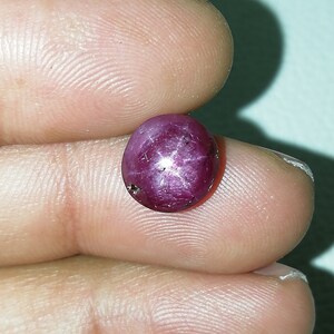 Könnte beinhalten: Ein polierter, ovaler Rubin-Edelstein. Der Edelstein hat eine tiefrote bis violette Farbe mit einem Sechs-Strahl-Stern-Effekt, bekannt als Asterismus, der auf seiner Oberfläche sichtbar ist. Der Rubin wird zwischen Fingern gehalten, um seine Größe und Farbe zu zeigen.