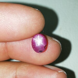 Könnte beinhalten: Ein ovalförmiger Rubin-Edelstein mit Sterneneffekt. Der Edelstein hat eine tiefrosa-rote Farbe, mit einem sechsstrahligen Sternmuster auf der Oberfläche. Der Rubin wird zwischen zwei Fingern gehalten.